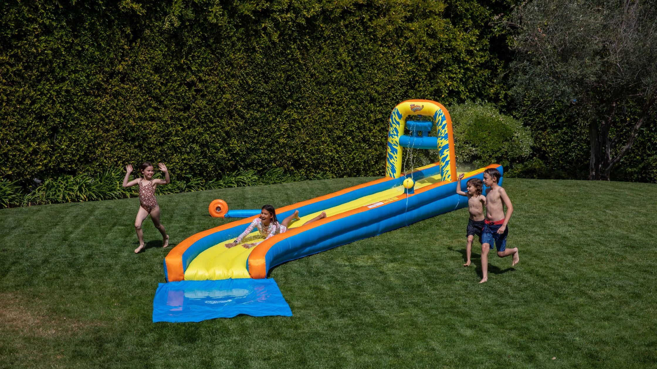Kids playing on inflatable backyard water slide with splash zone on green lawn
