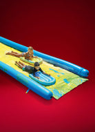 Two kids sliding on a yellow and blue inflatable slip and slide water toy against a red background